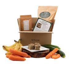 a box containing a Fleetful homecooking kit for homemade dog food, with fresh fruit and vegetables in front of it