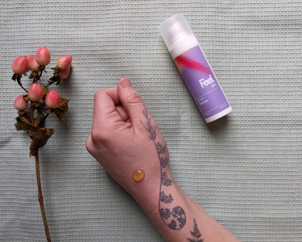 A tattooed arm with a blob of Feel's serum on, next to a bottle of the product and a sprig of berries