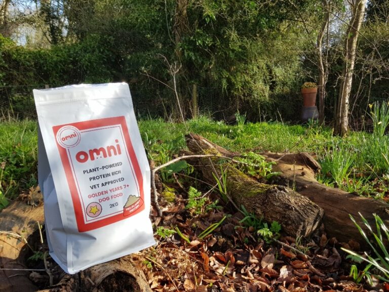 Bag of Omni dog food on a log surrounded by autumn leaves and with a background of trees and grass