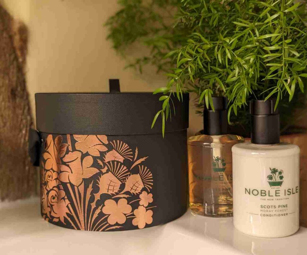 A beautifully designed cylindrical dark brown gift box with copper coloured flower patterns on, next to bottles of Noble Isle vegan shampoo and conditioner, in a bathroom setting.
