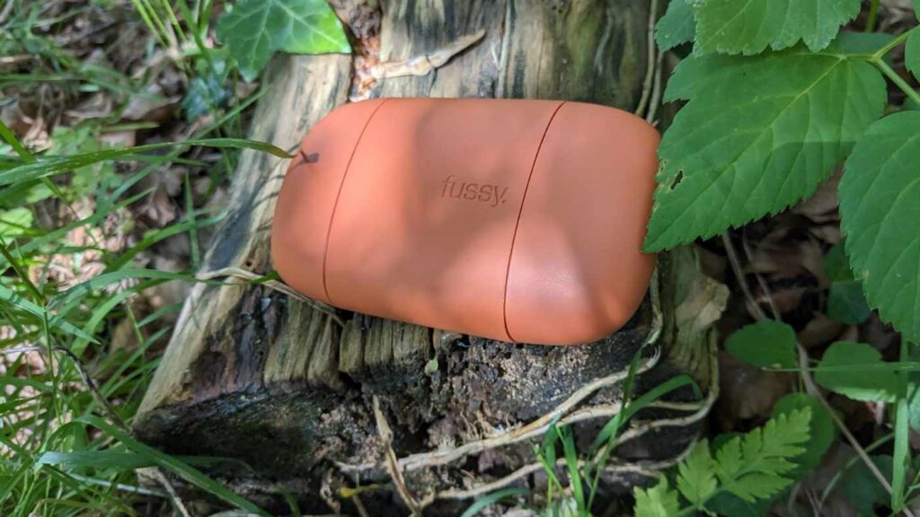 Orange plastic Fussy deodorant case nestled on a log in dappled sunlight