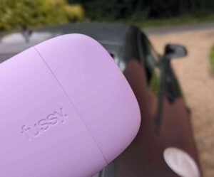 Plastic lilac coloured Fussy deodorant case in the foreground of the picture poking into the picture from the bottom left hand corner. Behind it is an out of focus dark grey car on a gravel drive