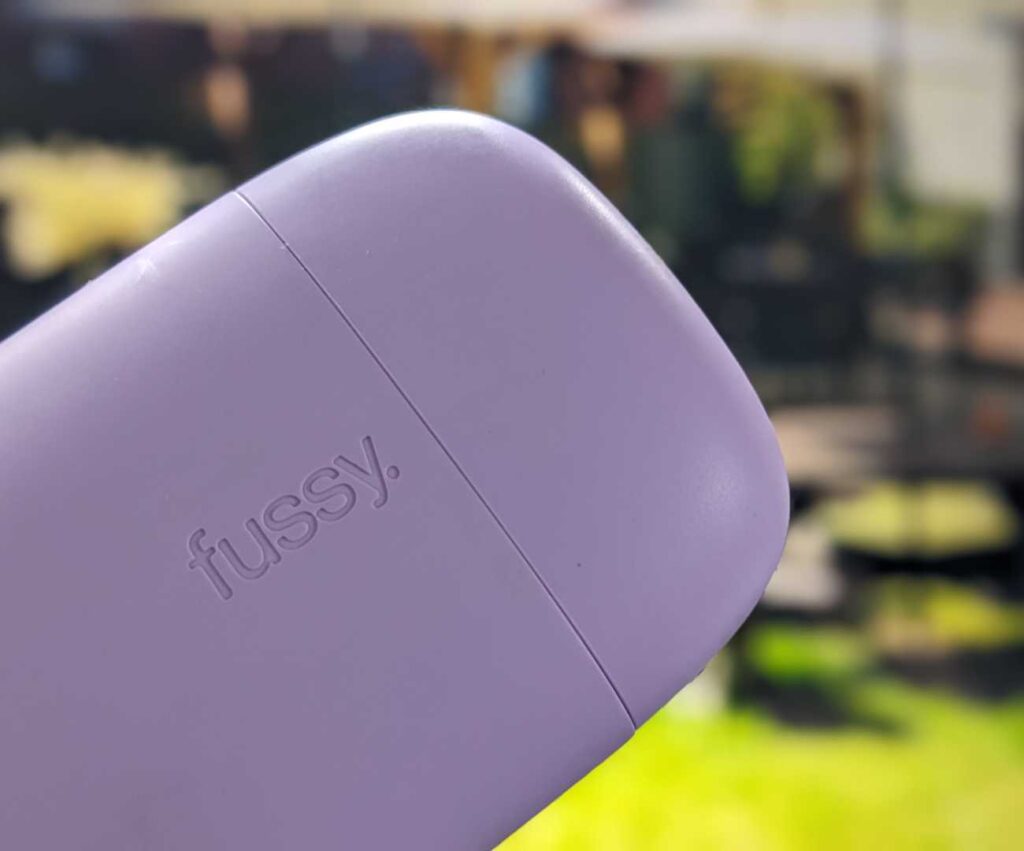 plastic lilac coloured Fussy deodorant case held in front of sunny pub garden in the background