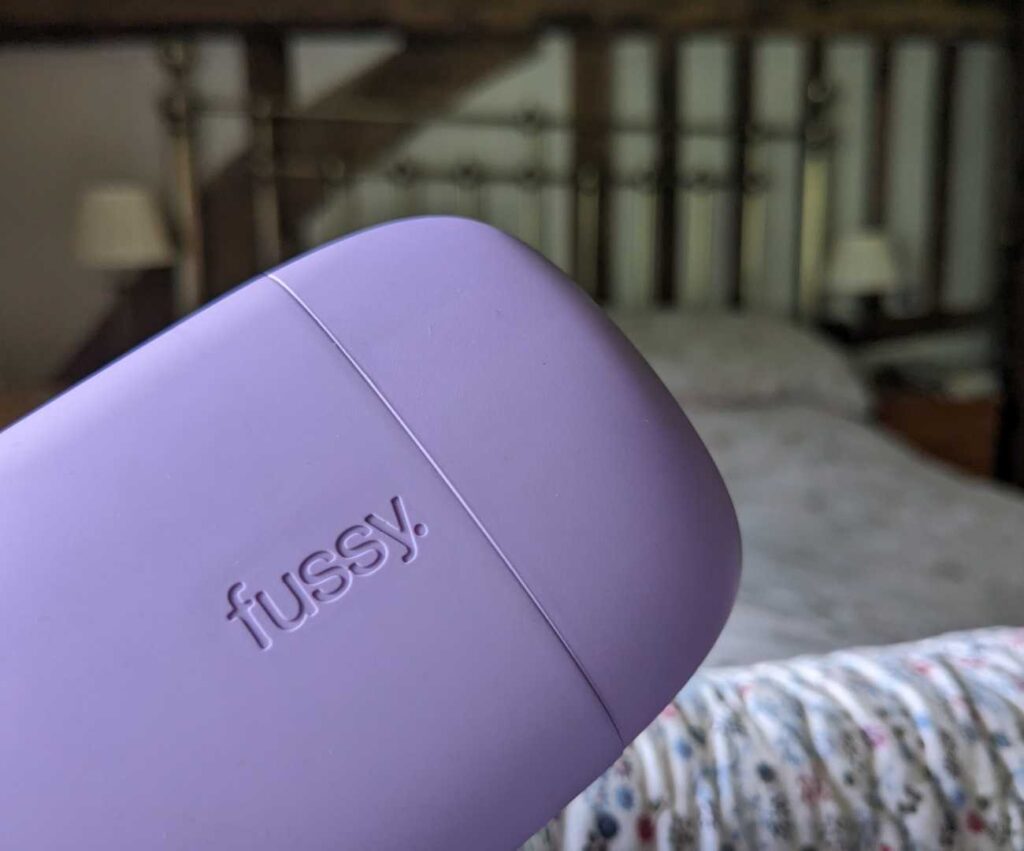 Plastic lilac coloured Fussy Deodorant case in a bedroom setting in front of an out of focus brass bed