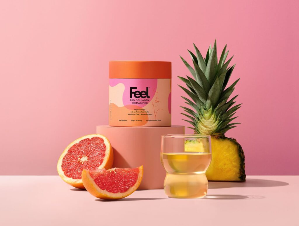 tub of feel pineapple pro collagen powder with a cut pineapple and segments of grapefruit each side. Set against a pink background with a glass of the diluted product in the foreground