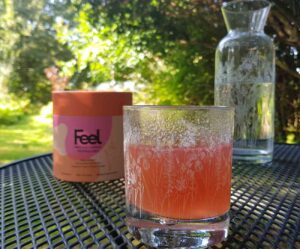 glass of Feel pineapple pro collagen diluted with water on a table in a sunlit garden under the trees. In front of a tub of Feel collagen and a carafe of water