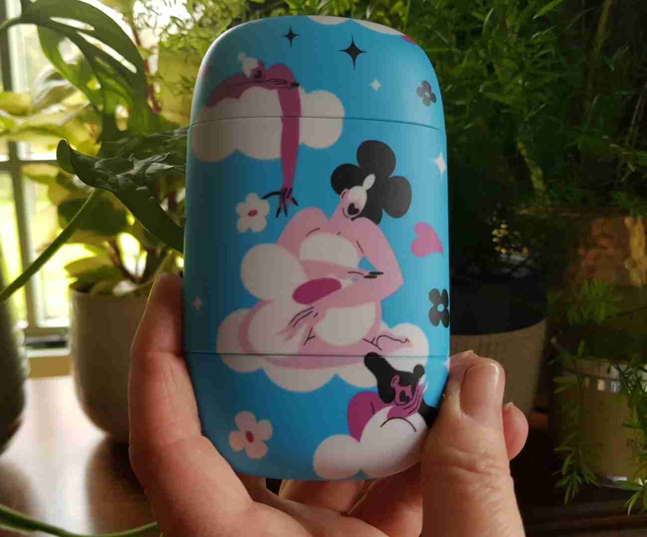 Finger and thumb of a hand holding a sky blue Fussy refillable plastic deodorant case, decorated with images of female caricatures sitting on clouds, with flowers dotted around in between. This case is supposed to represent Fussy deodorant's support for charities surrounding females and their periods. I am using this image to illustrate this Fussy deodorant review