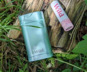 Pale green metallic Wild deodorant case, with the Wild logo at the bottom of the case. Next to the case is a small pink and white cardboard tube containing a travel sized Wild deodorant. The Wild logo is picked out on the white cardboard tube in large pink lettering