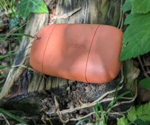 Rectangular burnt orange plastic Fussy deodorant case nestled on a log in dappled sunlight. The case has the brand name 'fussy' engraved into the front of the case. The log is surrounded by ivy leaves and blades of grass