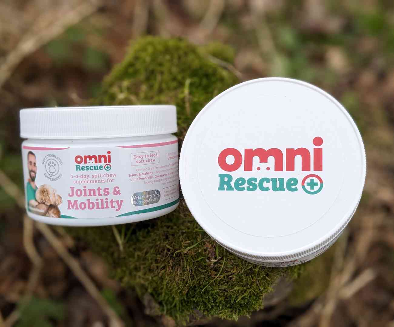 2 white plastic tubs resting on a moss covered tree stump. Both contain Omni dog supplements and are branded with the logo 'Omni Rescue'. 1 of the tubs is lying on its side, showing the logo and contents clearly, the other is with the top of the lid facing upwards, with the Omni Rescue branding in large letters