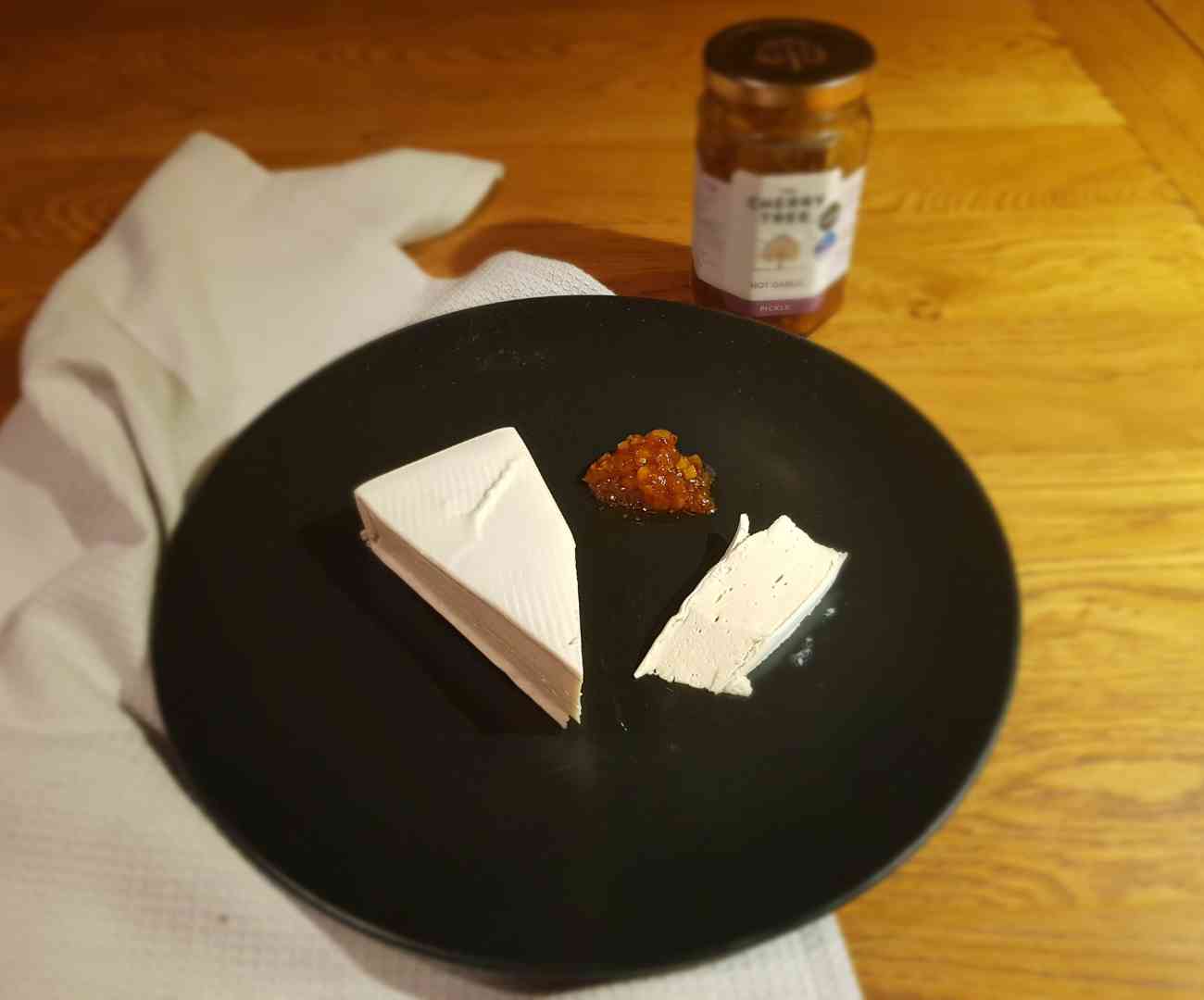 To illustrate this vegans and cholesterol article, this is an image of a black plate on a wooden table, with a wedge of vegan cheese, a thin slice next to it, and a small pile of brown-coloured pickle. The vegan cheese resembles a wedge of dairy brie. Behind the plate on the table is an indistinguishable glass jar with a white label on the front containing the pickle.
