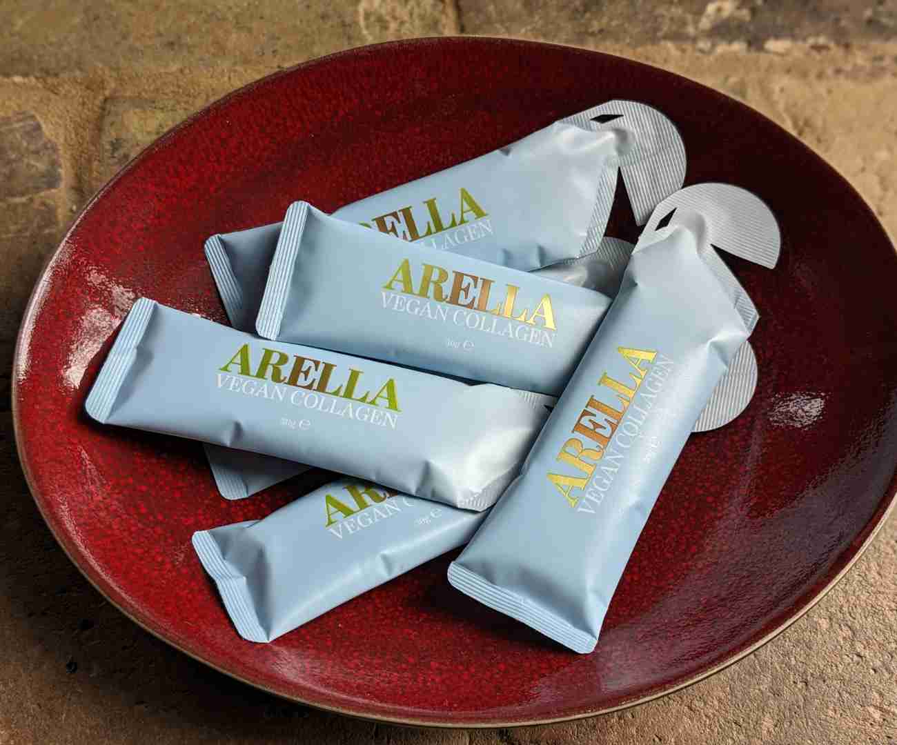 6 pale blue sachets of Arella vegan liquid collagen with gold and white lettering depicting the brand and the contents. The sachets are scattered in a deep red, oval ceramic bowl which is standing on a sand coloured brick floor