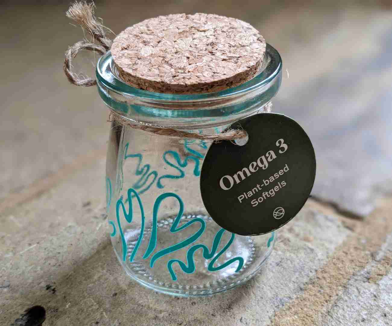 Image of a small clear glass jar, with blue/green swirly patterns painted on it, with a piece of string tied around the neck and a cork lid, giving it a rustic, natural feel. The jar is empty but is designed to contain soft gel capsules containing omega 3. The jar is sitting on a rustic brick floor. This is to illustrate this best vegan omega 3 supplements uk guide