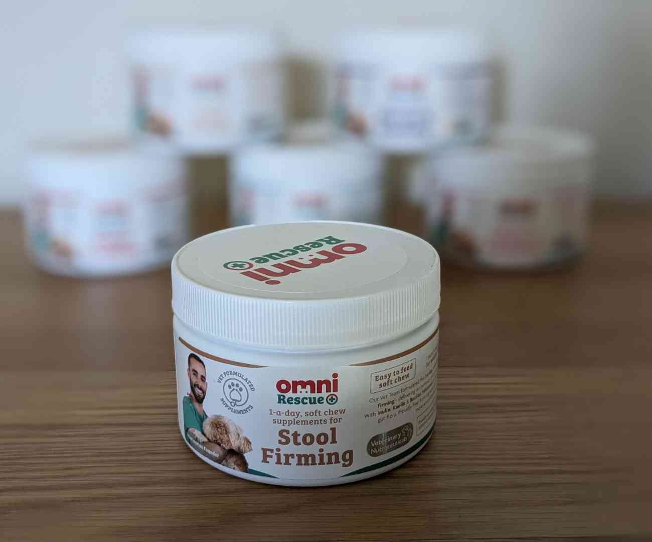 white plastic tub in the foreground, branded with 'Omni Rescue' logo, a picture of a vet holding a dog, and lettering on the front of the tub showing the contents - 1 a day soft chew Omni stool firming supplement for dogs. Additional lettering advises that the product is vet formulated. In the blurred background are 5 more stacked tubs of Omni supplements for different conditions